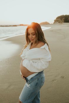 a pregnant woman standing on the beach at sunset with her belly exposed and hands in her pockets