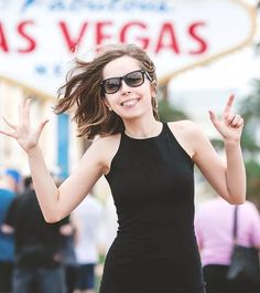 a woman in black dress and sunglasses making the peace sign with her hands while standing outside