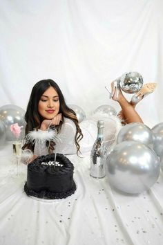 a woman sitting at a table with silver balloons and champagne in front of her holding a cake