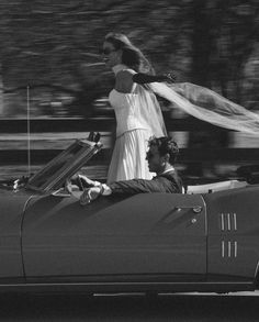 a woman riding in the back of a convertible car with a man standing next to her