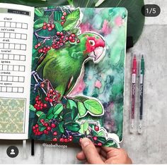 a hand holding an open notebook with a green bird on it and red berries in the background