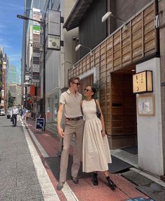 a man and woman are standing on the sidewalk in front of a building, posing for a photo