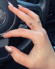 a woman's hand with a small cross tattoo on her left index finger next to a car steering wheel