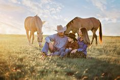 three people sitting in the grass with two horses