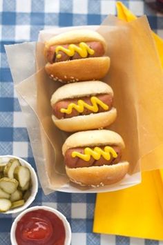 three hot dogs with mustard and ketchup in buns next to pickles