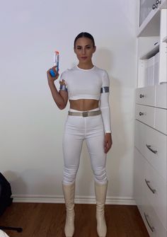 a woman standing in a room holding a toothbrush and wearing tight white pants with knee high boots