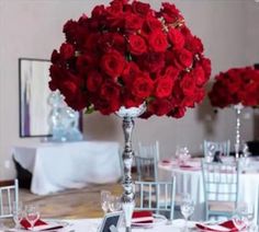 red roses in a silver vase on top of a table with white linens and place settings