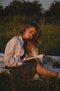 a woman sitting in the grass reading a book with her hair blowing back and eyes closed