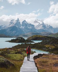 a woman walking down a wooden walkway towards mountains and water with snow capped peaks in the distance