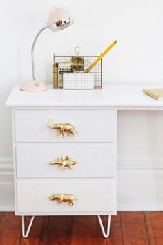 a white desk with three drawers and a lamp on top of it, in front of a white wall