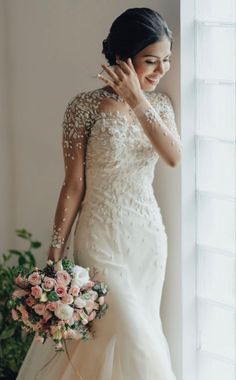 a woman standing next to a window in a wedding dress and holding a bridal bouquet