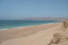 a sandy beach next to the ocean with people walking on it and mountains in the distance