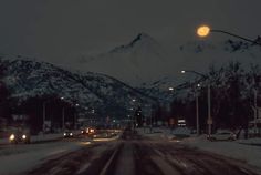 a snowy road with cars driving on it and the moon in the sky above mountains