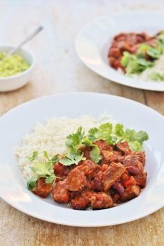 two white plates filled with meat and rice on top of a wooden table next to a bowl of guacamole