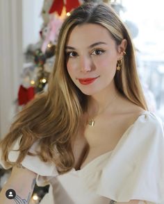 a woman with long hair wearing a white dress and red lipstick in front of a christmas tree