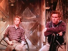 two young men sitting on chairs in front of a barn door, one looking at the camera