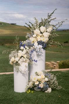 three white vases with flowers and greenery in them on the grass near an open field