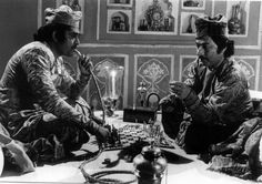 two men sitting at a table with chess pieces in front of them, one man is looking at the other