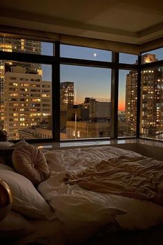 an unmade bed in front of a large window overlooking the city at night time