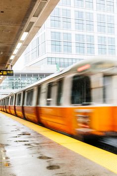 an orange and white train traveling down tracks next to a tall building with lots of windows