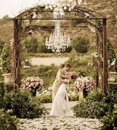 a bride and groom kissing under an outdoor wedding ceremony arch with chandelier in the background