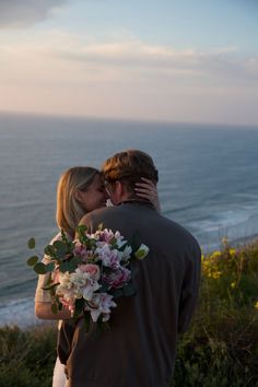 a man and woman standing next to each other on top of a hill near the ocean