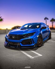 a blue car parked in a parking lot next to palm trees at sunset with the sun setting