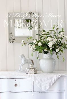 a white vase with flowers sitting on top of a dresser next to a mirror and a small figurine