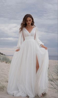 a woman standing on top of a sandy beach wearing a white dress and long sleeves