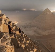 two people are sitting on the edge of a cliff with pyramids in the background