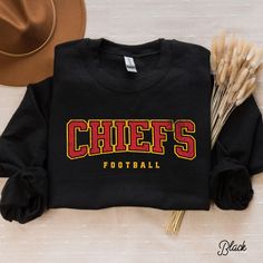 a black shirt with the word chiefs on it next to a hat and some dry grass