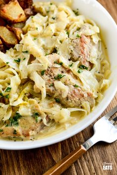 a white bowl filled with pasta and meat on top of a wooden table next to a fork