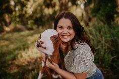 a woman holding a baby goat in her arms and smiling at the camera with trees behind her