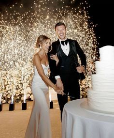 a man and woman standing next to each other in front of a wedding cake with fireworks behind them