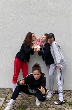 three women pose for a photo with their dolls in front of a white wall and cobblestone walkway