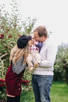 a man and woman holding a baby in front of an apple tree