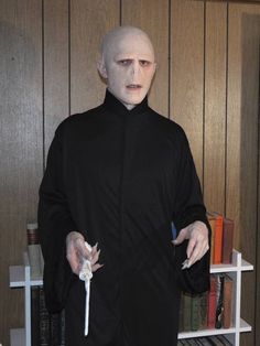 a man dressed in black with white makeup and fake hands holding something while standing next to a bookshelf