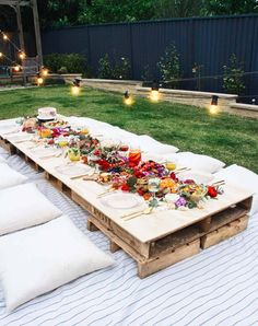 a long table is set up outside on the grass with pillows and lights around it