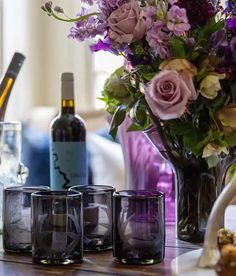 three wine glasses sitting next to each other on a table with flowers and bottles in the background