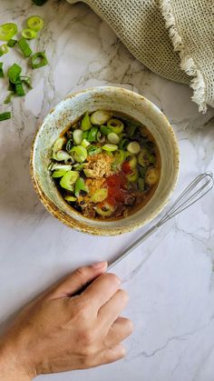 a hand holding a spoon in a bowl filled with soup on top of a marble counter