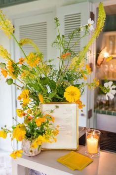 yellow flowers and greenery in a vase on a table next to a lit candle