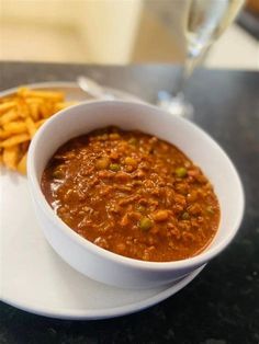 a white plate topped with a bowl of chili and french fries next to a glass of wine