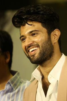 a man with a beard smiling and wearing a tan vest over a white shirt in front of two men