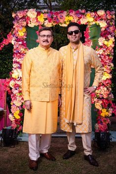 two men standing next to each other in front of a flower covered arch with pink and yellow flowers