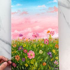 someone is painting flowers in the grass with pink and blue sky behind them on a canvas