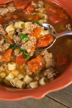 a red bowl filled with meat and vegetable soup on top of a wooden table next to a spoon