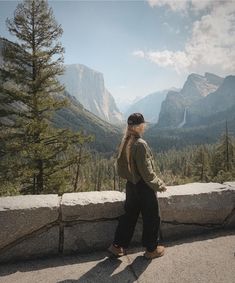 a woman standing on the edge of a cliff looking at trees and mountains in the distance