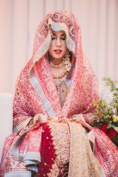 a woman in a red and white wedding outfit sitting on a chair with flowers behind her