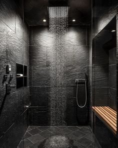 a shower head in the middle of a black tiled bathroom with wooden bench under it