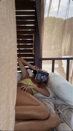 a woman laying on top of a bed with a book in her hand next to a window